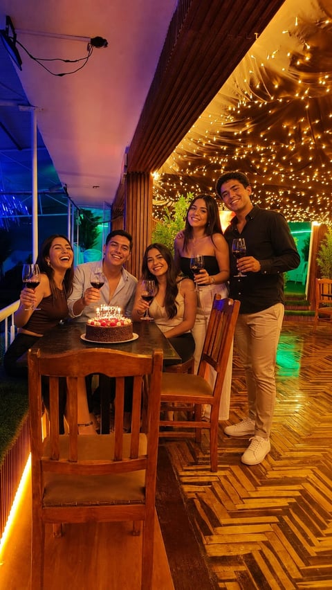 Group of five people celebrating with drinks and a birthday cake on a decorated outdoor terrace with string lights and colorful ambient lighting