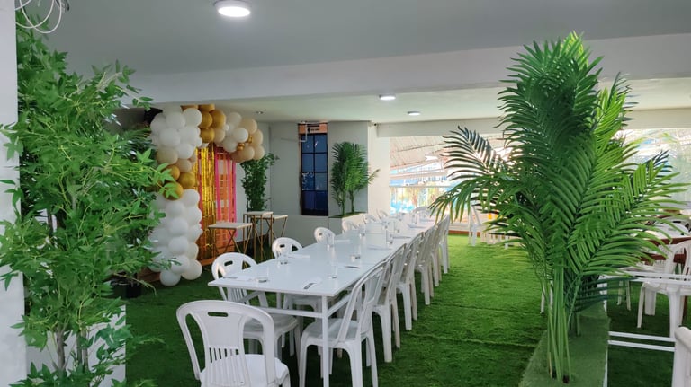 Salón con sillas blancas, mesas, grass artificial y plantas tropicales